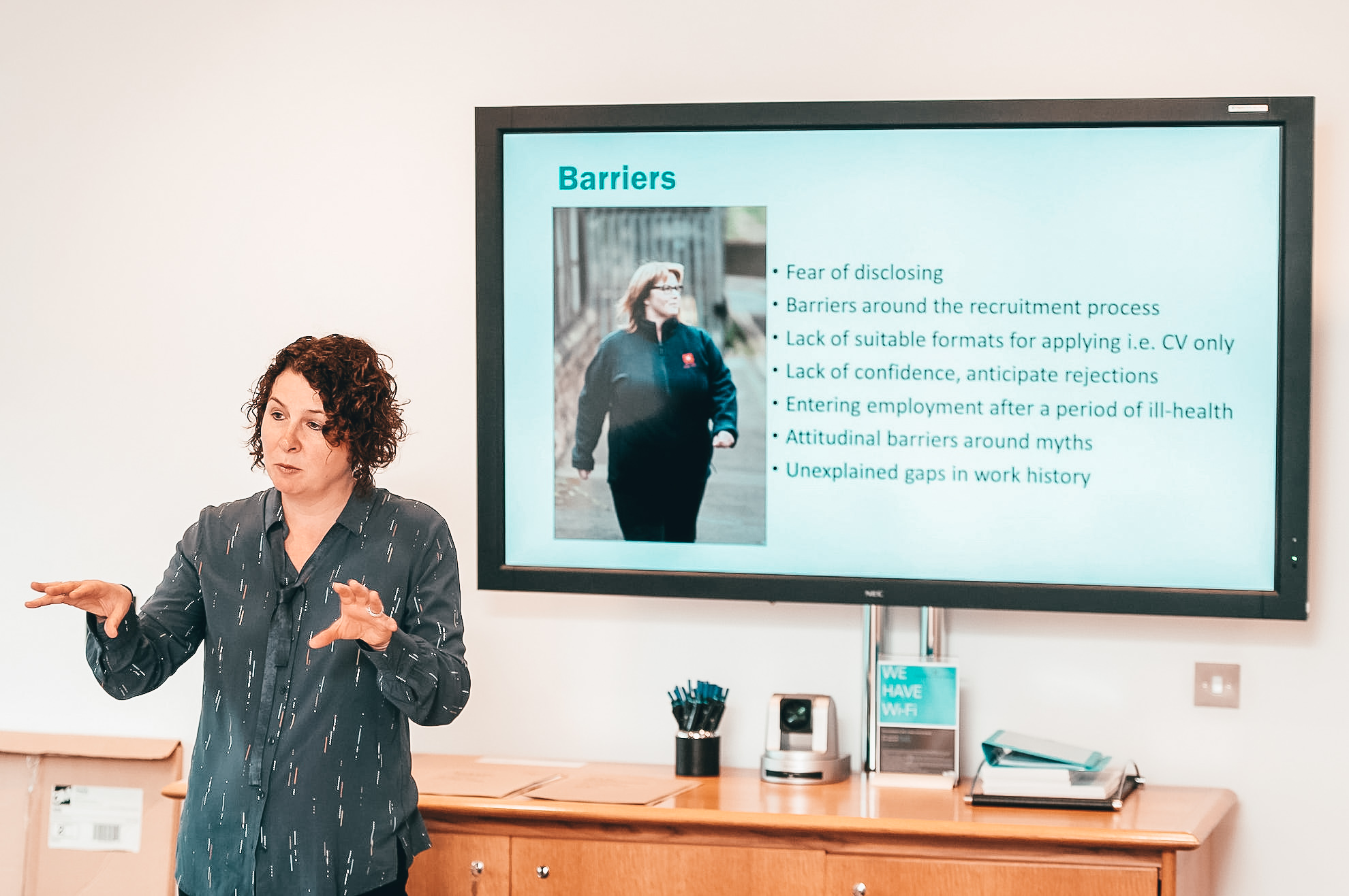 A woman presents a talk to a room, with a whiteboard on the wall behind displaying information under the heading 